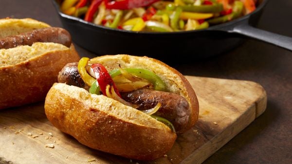 一个小香肠和胡椒三明治卷放在棕色表面的木制砧板上。后面放着一锅炒辣椒。