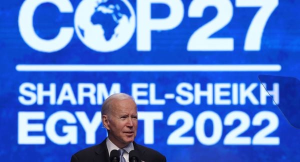 在COP27大会上，美国总统乔·拜登站在讲台上对着麦克风讲话，他身后的屏幕上显示着蓝白相间的会议标志和地点。