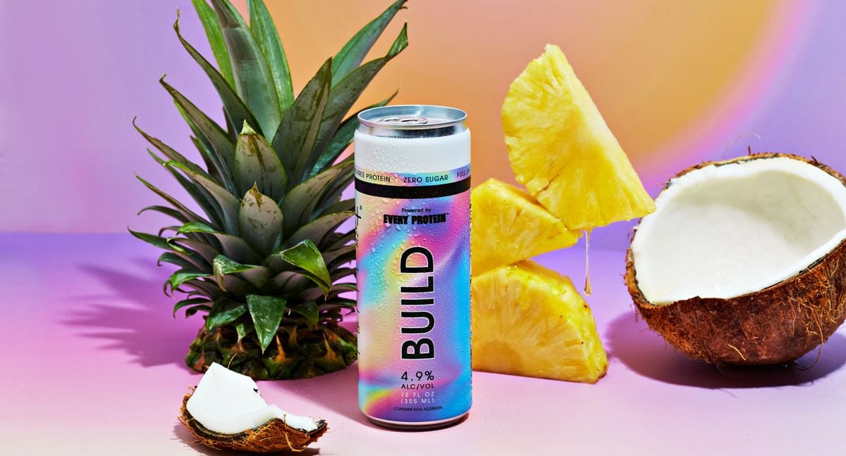 彩虹形状的Build可乐罐，旁边是菠萝顶部，粉彩背景上是碎椰子片。