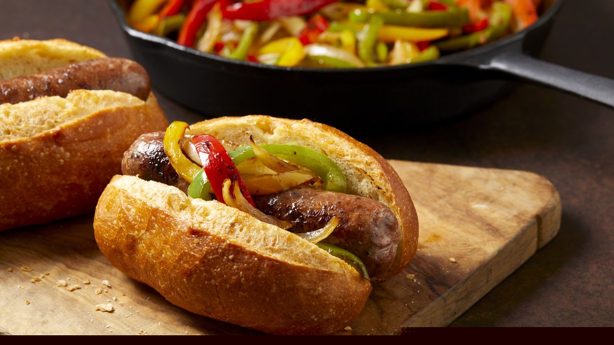 一个小香肠和胡椒三明治卷放在棕色表面的木制砧板上。后面放着一锅炒辣椒。