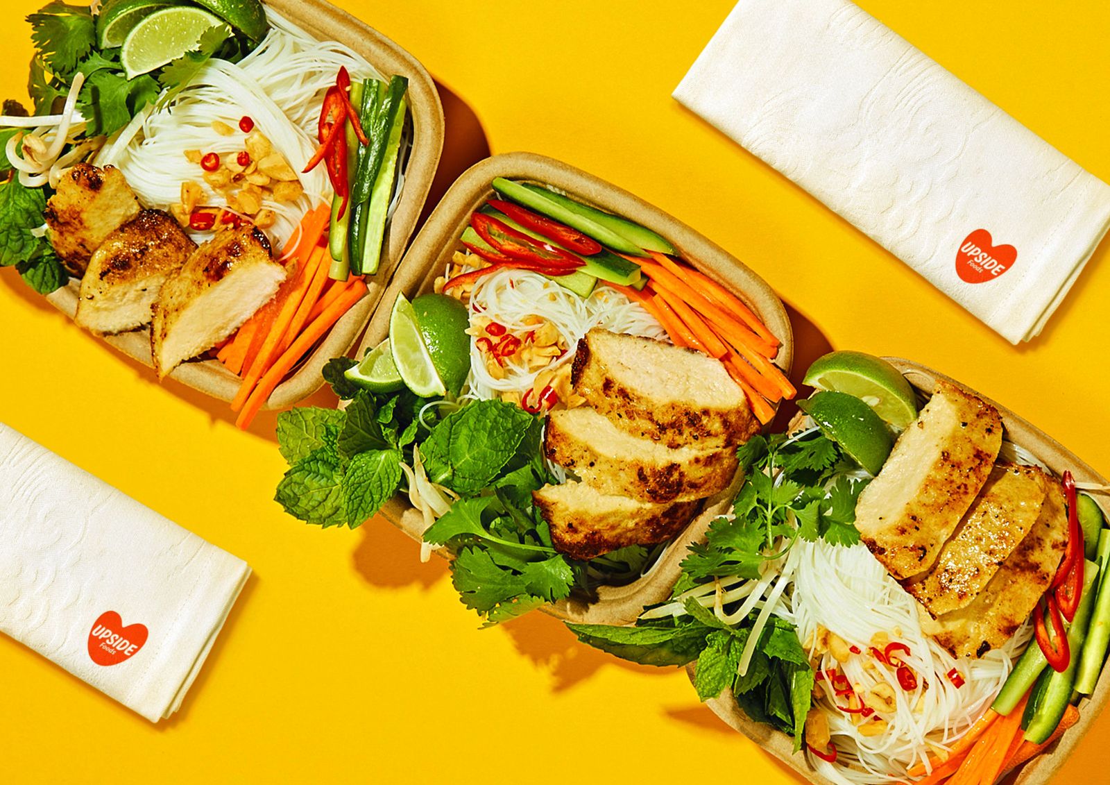 三个方形的纸容器里装着鸡肉块、蔬菜和蔬菜，旁边还有黄色背景的Upside Foods餐巾