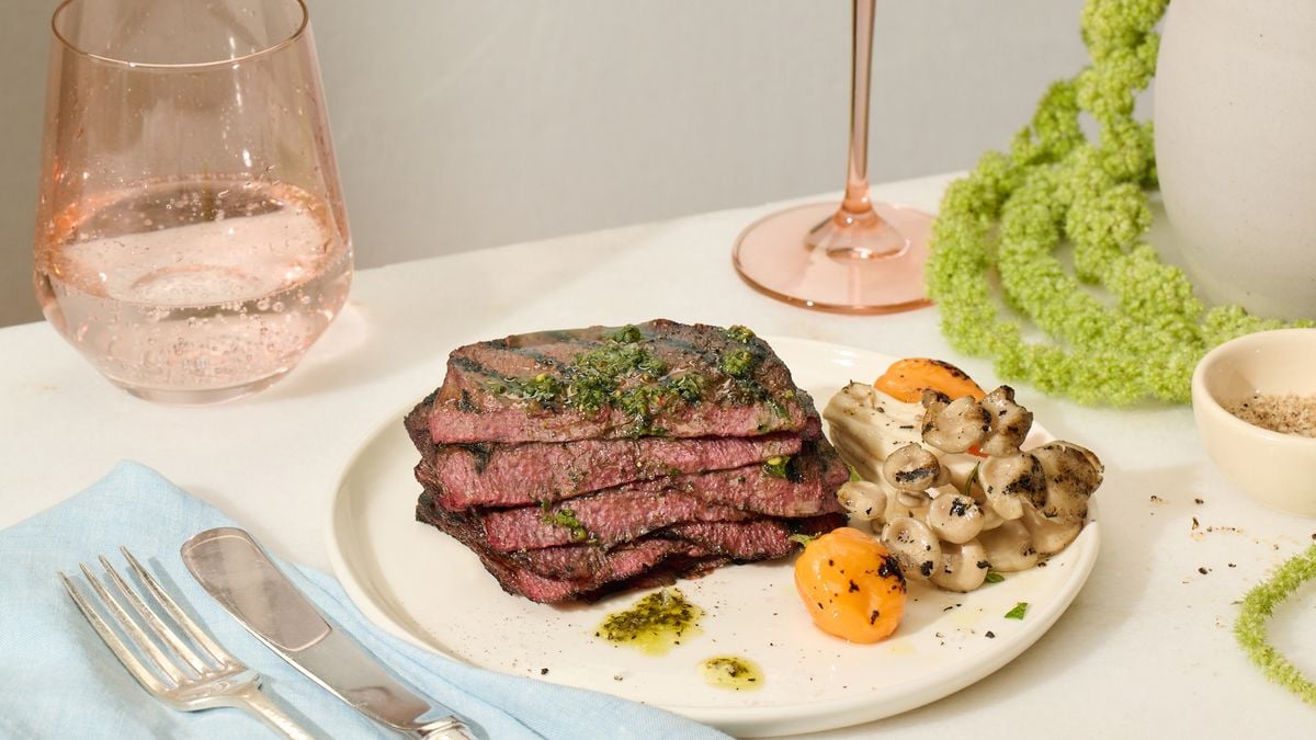 A white plate with a stack of five grilled thin steaks with a green dressing on top. The plate also has mushrooms and small grilled peppers. The plate is on a table set for dinner.