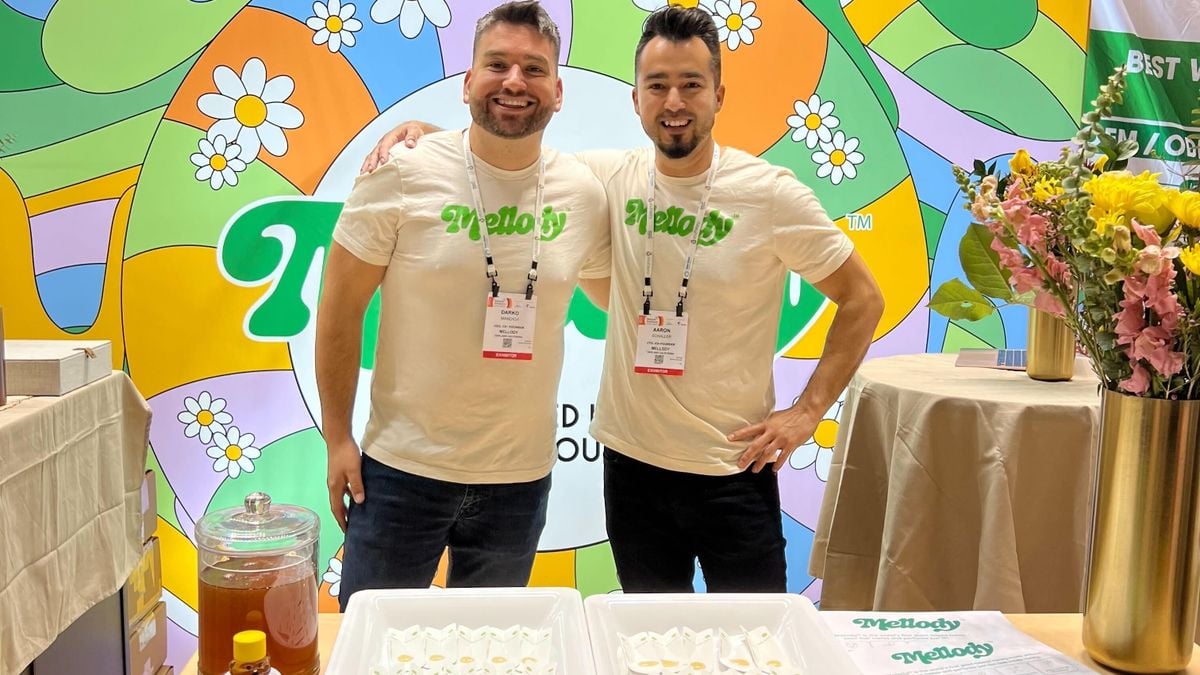 达科·曼迪奇(Darko Mandich)和亚伦·夏勒(Aaron Schaller)站在Mellody品牌的一个彩色摊位前，摊位上有品牌背景，他们面前的桌子上放着蜂蜜试勺。