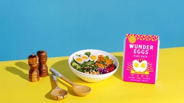 A white bowl with a salad that has three WunderEgg halves sits on a yellow surface. A pink-labeled package of WunderEggs is at right, and wooden salad tongs and salt and pepper shakers are at left