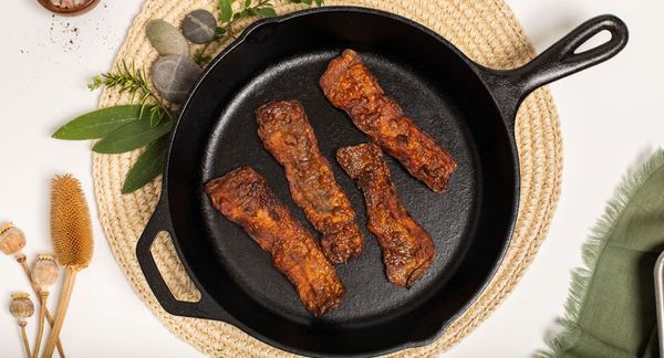 一个铸铁煎锅里放着四片煮熟的“我的培根”，放在米色圆形编织餐垫上，左上方放着香草和石头，还有一小盘粉红色的盐。