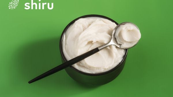 Overhead view of a bowl of Shiru's OleoPro fat with a spoon lying on top of it. The background is green and features Shiru's logo.