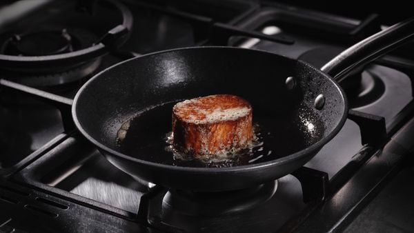 A black frying pan on a black gas range with a sizzling Juicy Marbles Thick Cut Filet cooking inside.