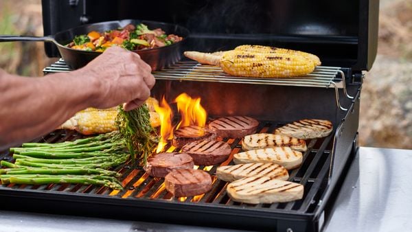 一只手拿着一捆香草，放在一个火热的双层烤架上，左下有烧烤痕迹的肉排和肉排，右下有芦笋，还有一锅炒菜和玉米