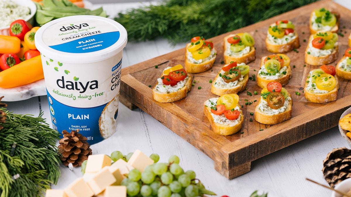 柜台上有一个木制的餐盘，上面放着涂着奶油奶酪的小面包片，上面放着切片的圣女果和香葱，旁边放着一桶Daiya奶油奶酪和其他东西。