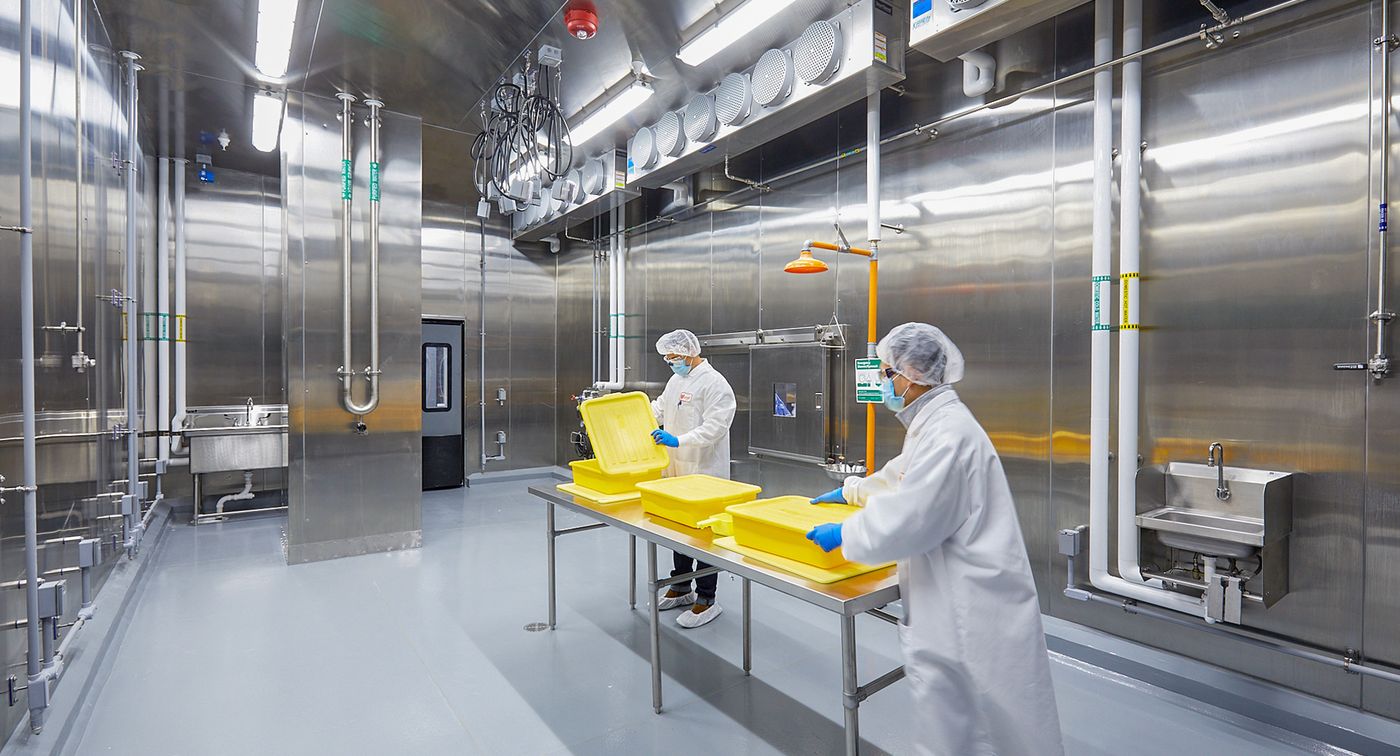 穿着食品安全防护装备的人们在一个有金属墙、管道和风扇的房间中间的金属桌子上拿着黄色的塑料桶。
