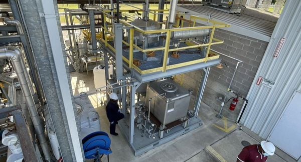 发酵设备在一个开放的建筑中，一面是混凝土砌块墙和地板，后面是一个开放的草地和树木的视野。两个戴安全帽的人站在附近。