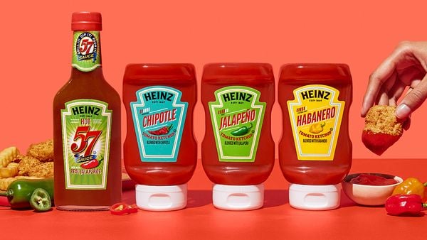一瓶57辣酱和三瓶亨氏辣酱放在深粉色背景的红色表面上。左边是墨西哥辣椒片，右边是一只手在蘸番茄酱