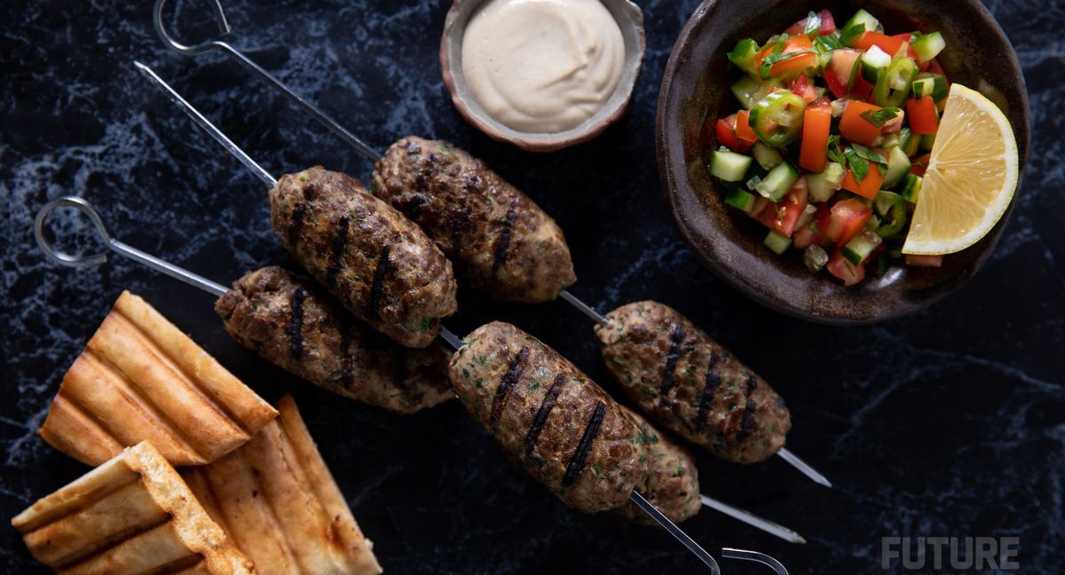 大理石表面上烤着烤好的羊肉串，一边是烤的三角皮塔饼，另一边是酸奶酱和番茄黄瓜胡椒蔬菜配菜。