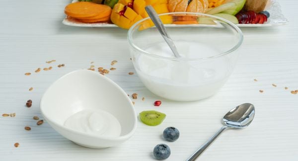 一小盘由细胞培养的奶制品制成的原味酸奶放在一个较大的酸奶玻璃碗前面。前面放着一把勺子，背景是切水果的砧板。